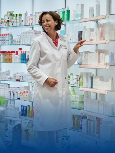 Eine Apothekerin in weißer Arbeitskleidung steht neben dem Medikamentenregal.